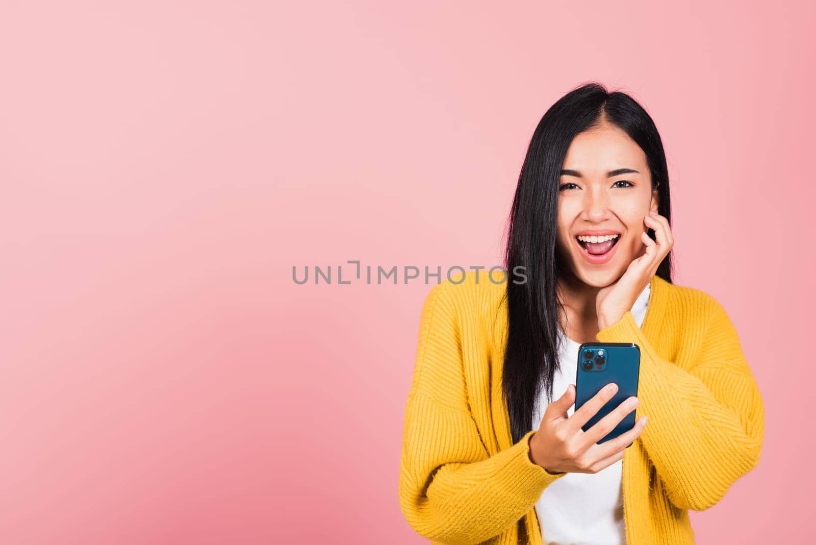 Happy Asian portrait beautiful cute young woman teen smiling excited using smart mobile phone studio shot isolated on pink background, Thai female surprised making winner gesture on smartphone