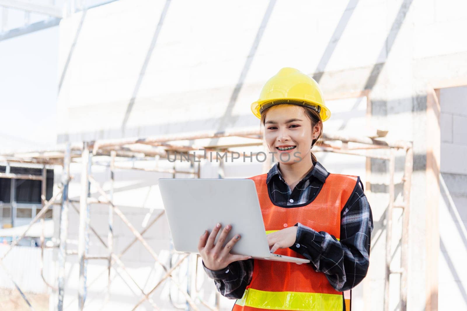 Asian engineer architect worker woman holding laptop inspect and oversee infrastructure progress at construction site, engineering use computer to operate project and control worker to building