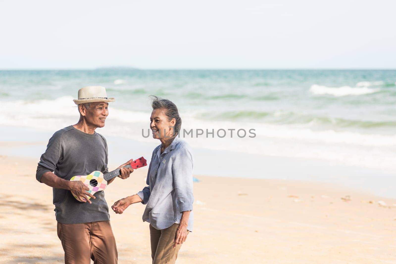 Happy senior couple relaxing outdoors singing and playing acoustic guitar at beach near sea sunny day, Mature man playing ukulele for his wife at sea, plan life insurance at retirement couple concept