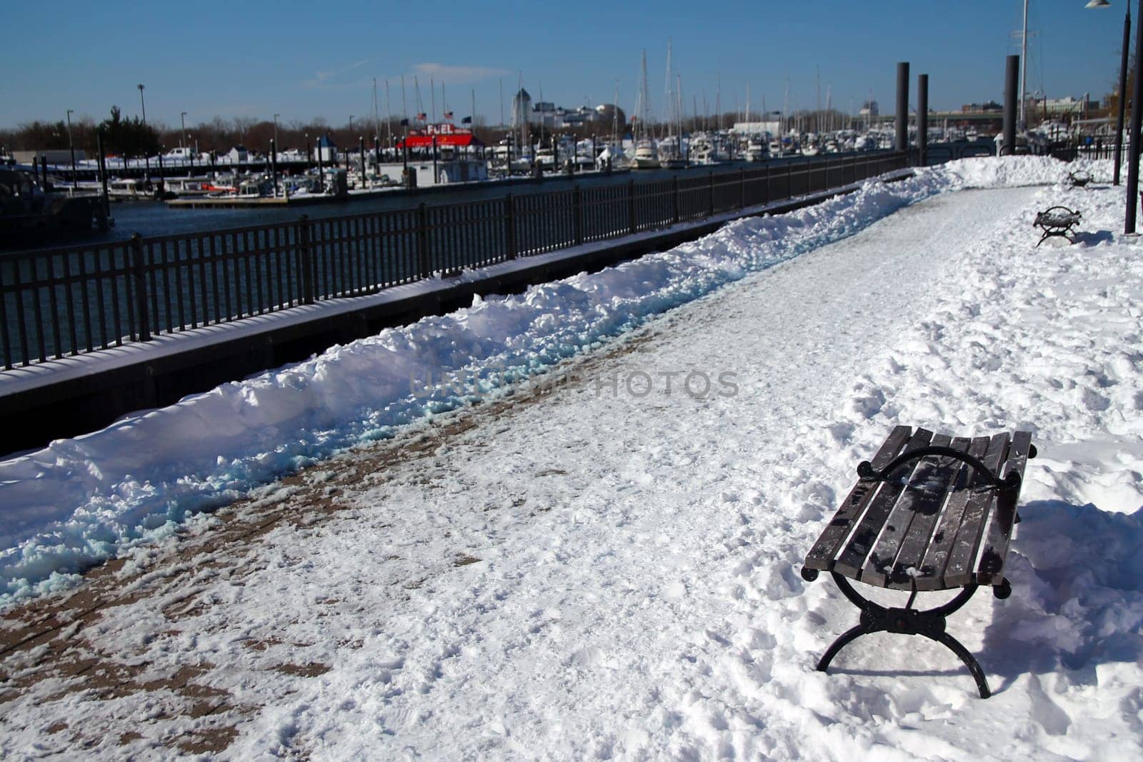 The path in the snow and the bench on the Morris Channel Basin Walkaway in Jersey City in a sunny winter day