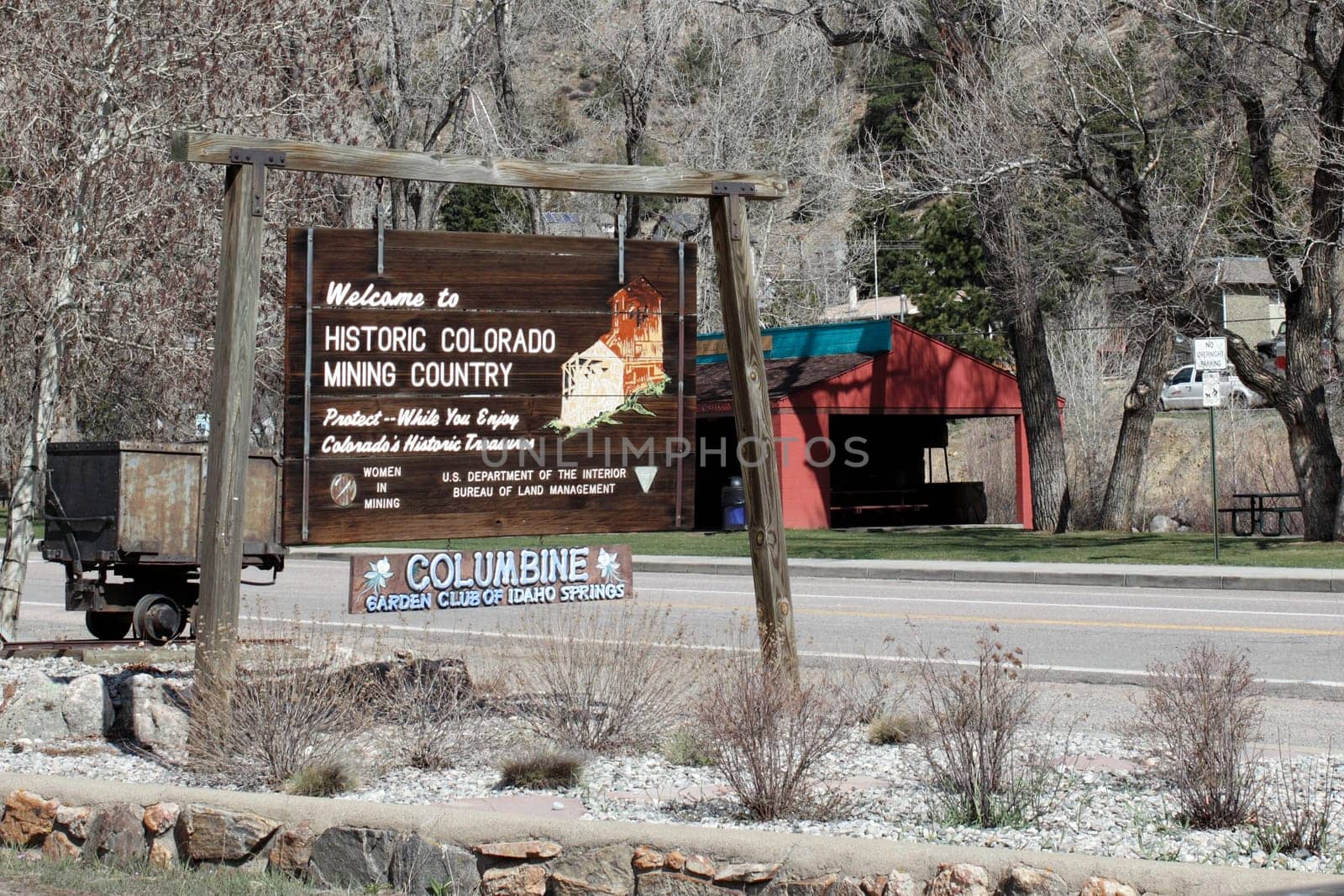Idaho Springs, CO, USA - April 21, 2014: Old wooden sign welcoming visitors to historic Colorado mining country with other information. The sign has a painting of the towns Argo Mine as well as an antique mining cart behind the sign on a sunny spring day.