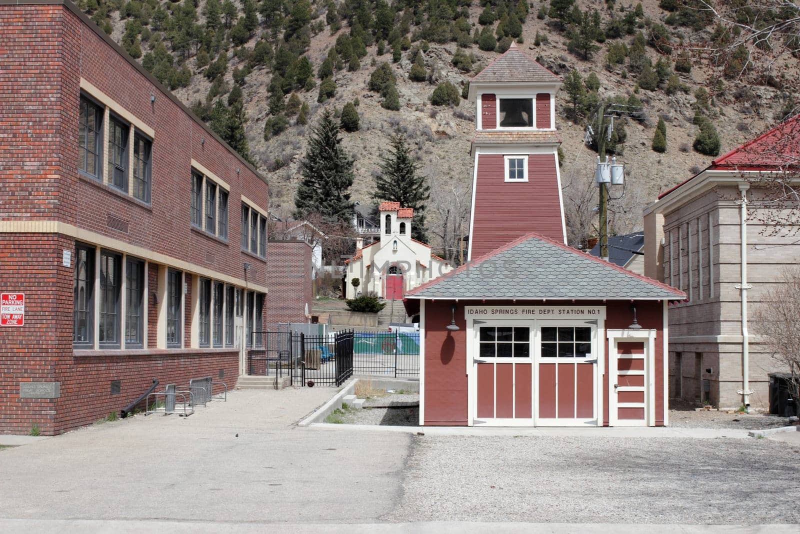 Idaho Springs, CO, USA - April 19, 2014: Historical architecture rises up with a bell on top of this old and small, reddish brown fire station located in the west part of downtown. A rare antique fire station with old fashioned doors next to a school and the main library, near a picturesque church all with various shades of red in front of a sunny mountain background.