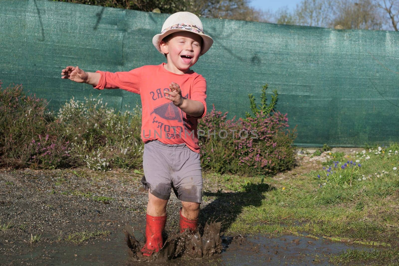 Happy male kid in rubber boots have fun playing in a puddle mud