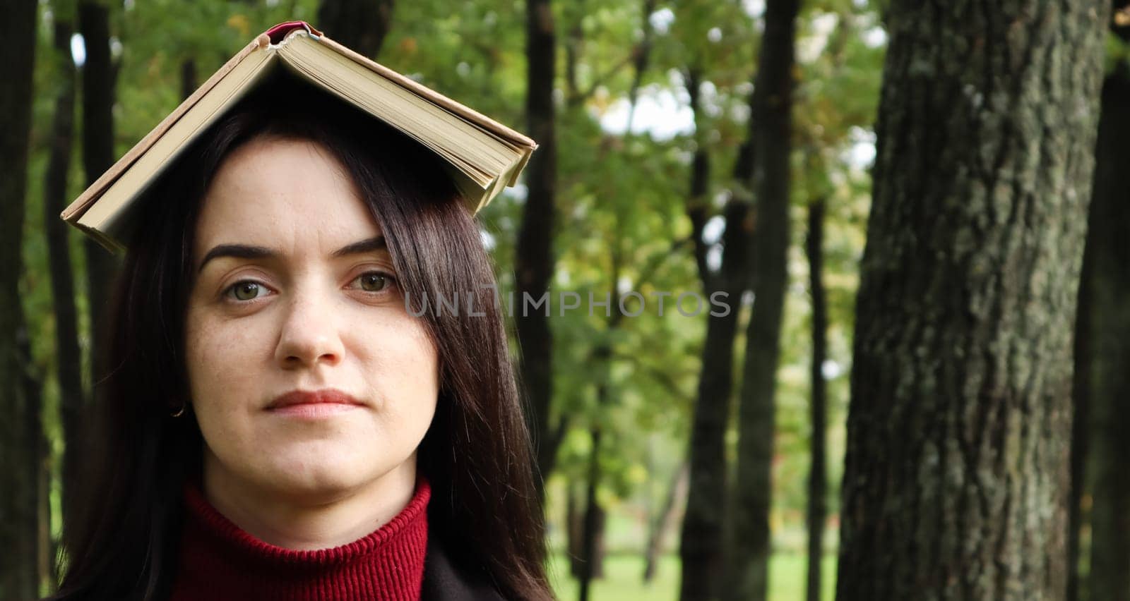 Portrait of a young and funny brunette in the park holding an open book on her head. Learning is fun. Woman balancing with books on her head. The student is tired of reading