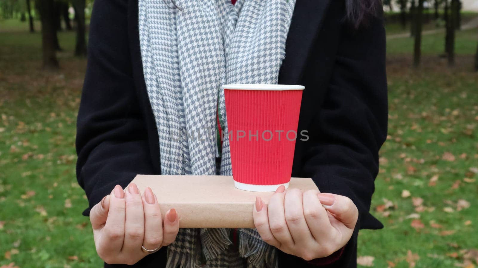 A young woman in a black coat and a gray scarf holds one book and a glass with coffee in her hands on the background of an autumn park. On open air. Education, close hands of the girl