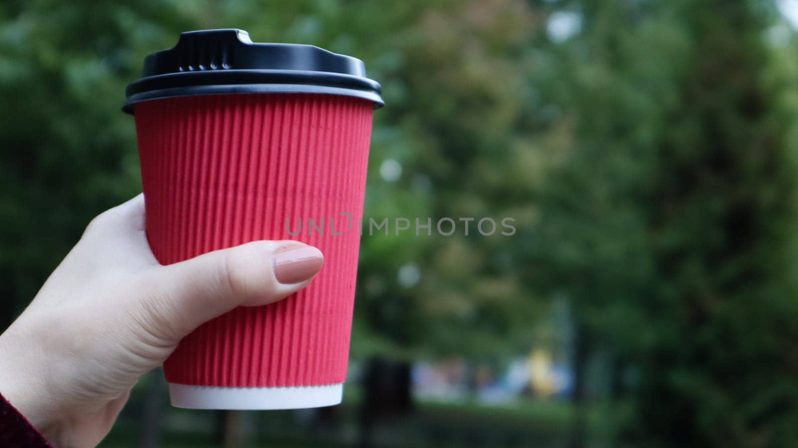 A young girl in a coat holds morning coffee with her while walking in the park. Hand holding paper cup of coffee in green park. Takeaway cappuccino. Close-up, copy space