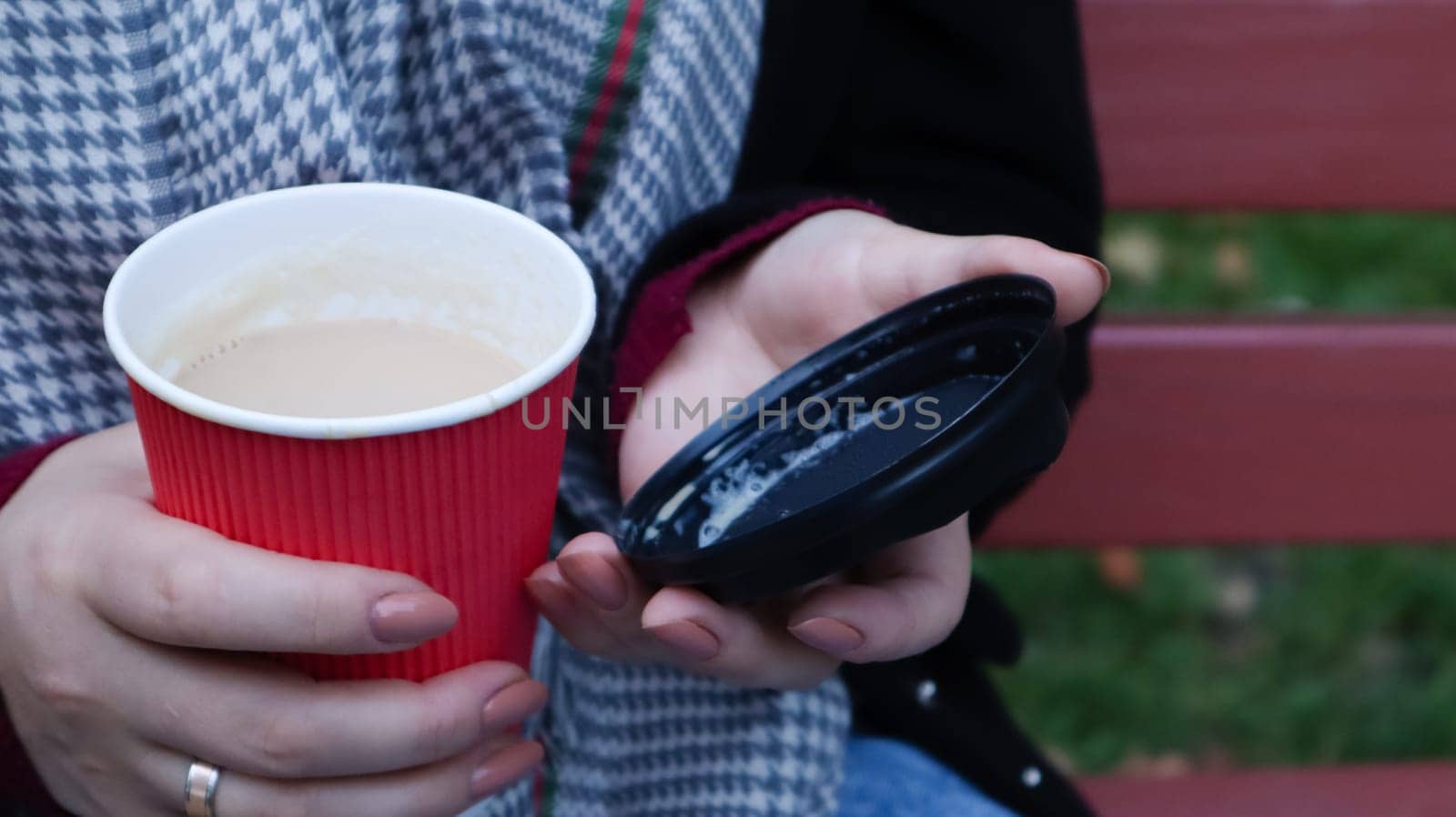 Female hands are holding morning takeaway coffee and closing a paper cup with a lid while sitting on a park bench, close-up of hands