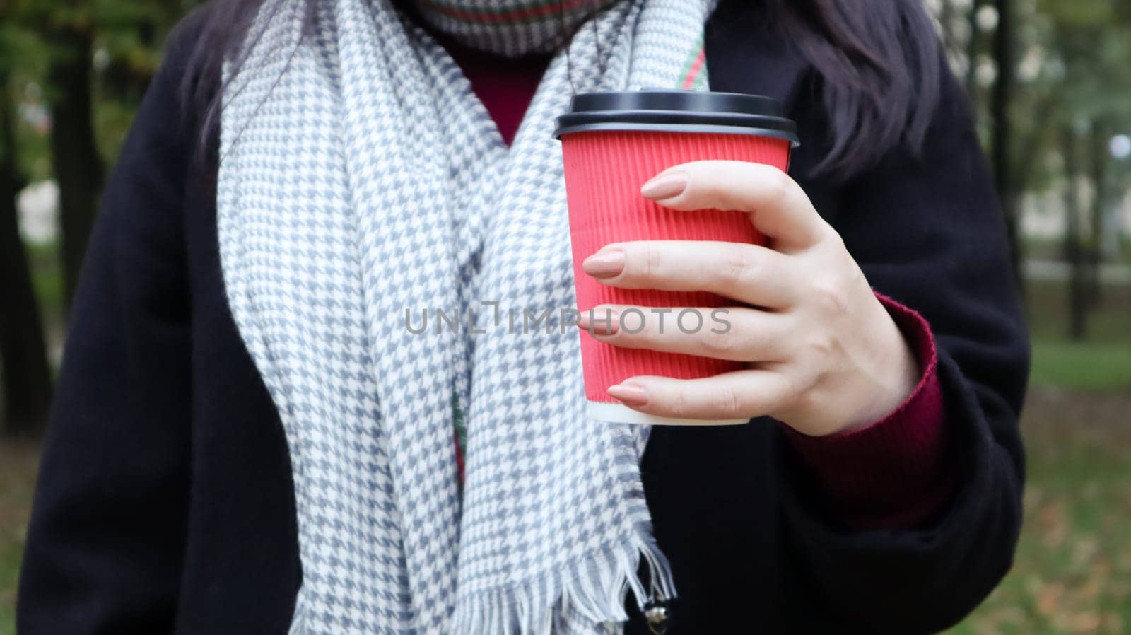 A young girl in a coat holds morning coffee with her while walking in the park. Hand holding paper cup of coffee in green park. Takeaway cappuccino. Close-up