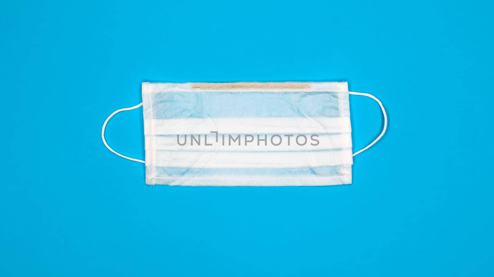 One 3-ply disposable surgical face mask with rubber grips covering mouth and nose against blue background. Protection from bacteria, healthcare and medicine concept. COVID-19. A dangerous virus