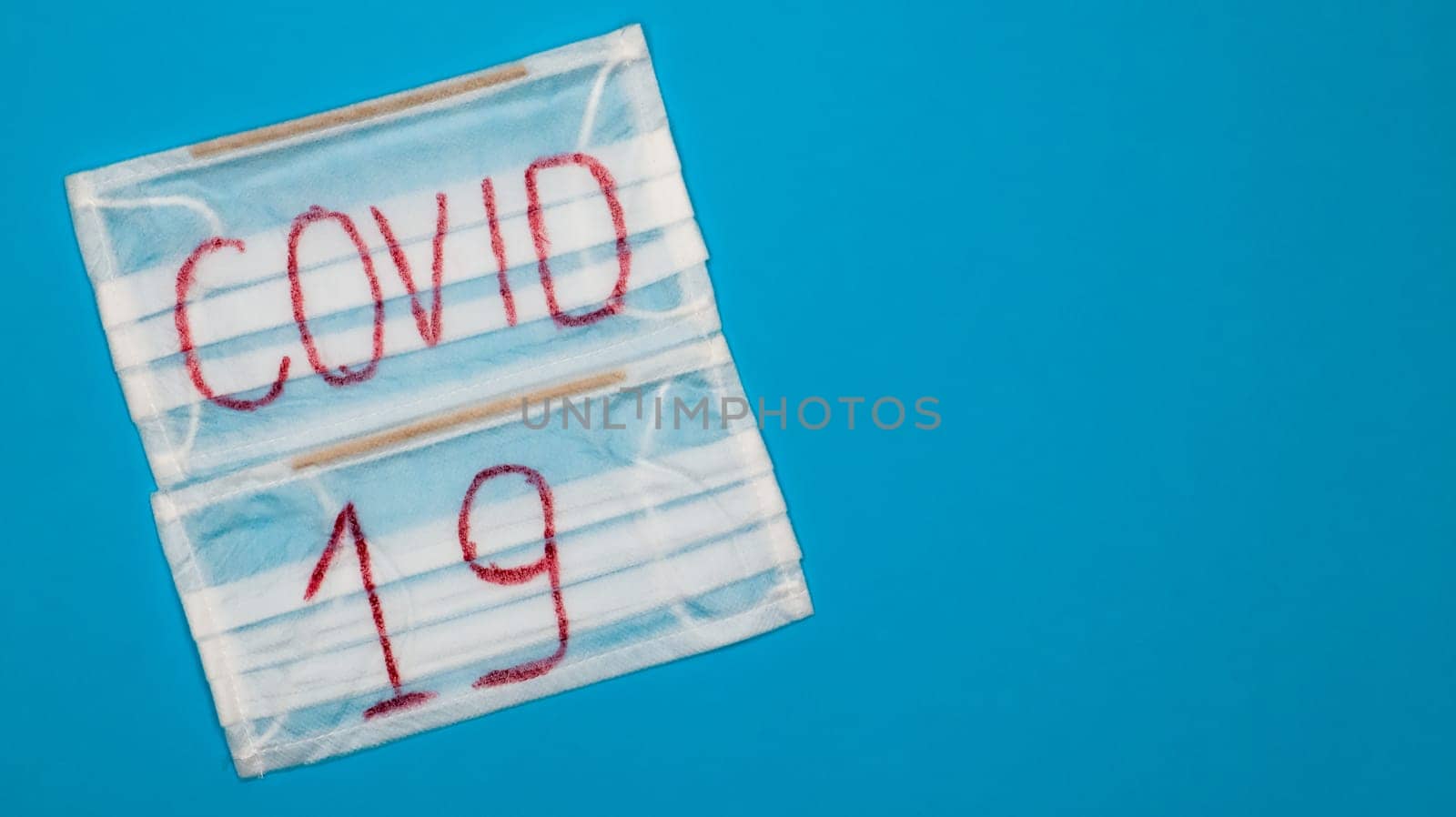 Typical white 3-ply surgical mask with rubber earhooks to cover mouth and nose with English block letters Covid-19 on a blue background. Dangerous virus and quarantine concept. Copy space.