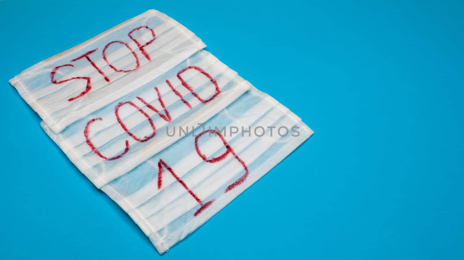 Typical 3-ply white surgical mask with rubber earhooks to cover mouth and nose with English block letters Stop Covid-19 on a blue background. Dangerous virus and quarantine concept. Copy space.