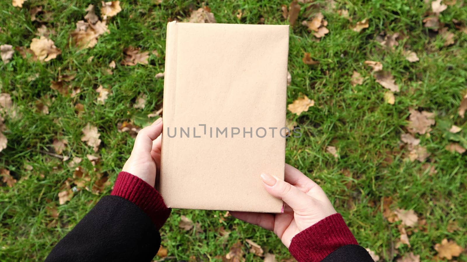 A closed book in a cover made of craft paper in female hands with green grass and fallen yellow leaves in the background. Flat lay, top view. Template, layout. Copy space