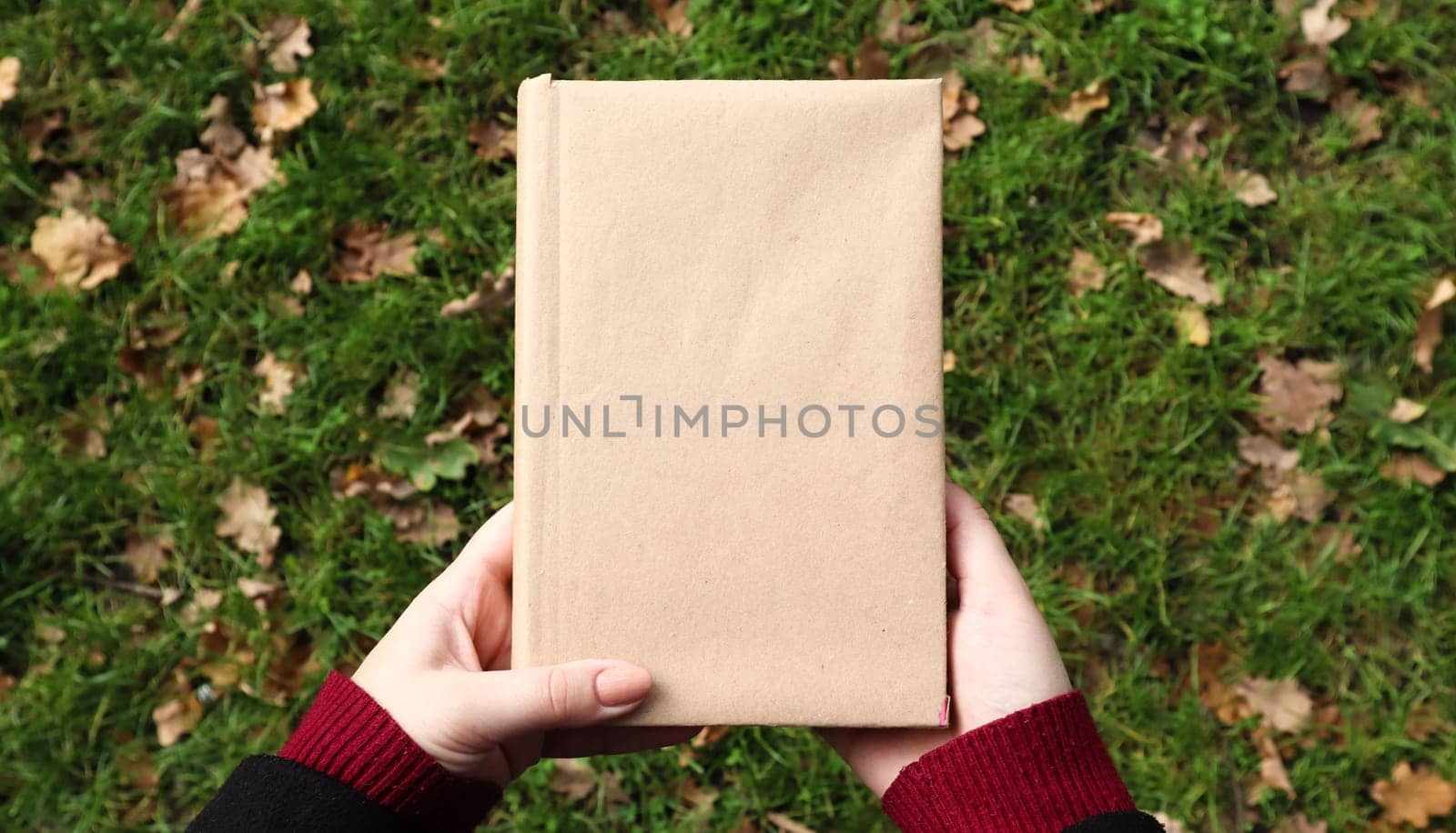 A closed book in a cover made of craft paper in female hands with green grass and fallen yellow leaves in the background. Flat lay, top view. Template, layout. Copy space