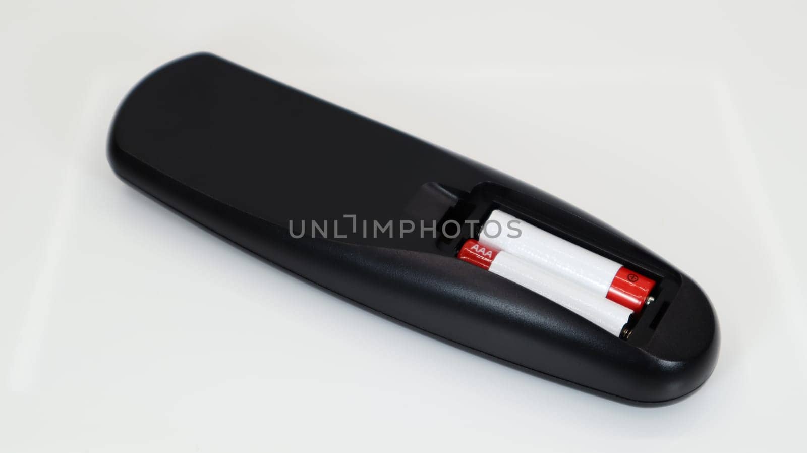 Black TV remote control with AAA alkaline batteries in red and white on a white background. Battery replacement, spare parts. Remote control battery compartment close-up
