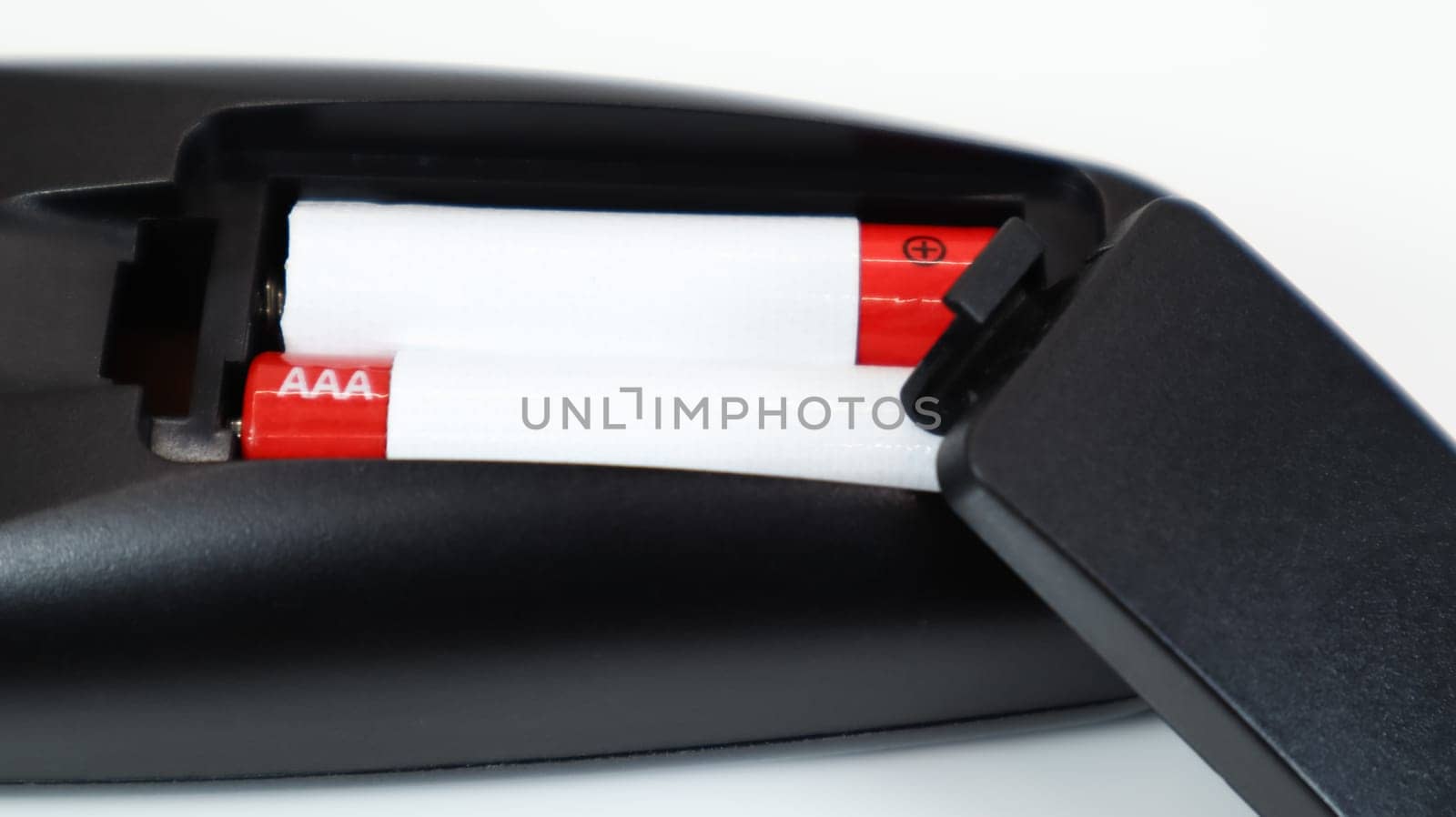 Black TV remote control with AAA alkaline batteries in red and white on a white background. Battery replacement, spare parts. Remote control battery compartment close-up