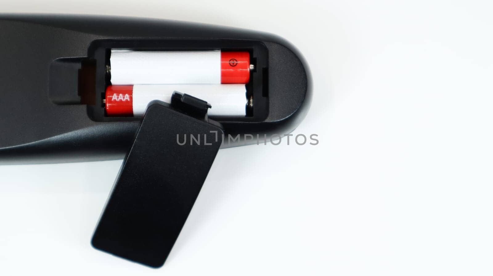 Black TV remote control with AAA alkaline batteries in red and white on a white background. Battery replacement, spare parts. Remote control battery compartment close-up. Copy space