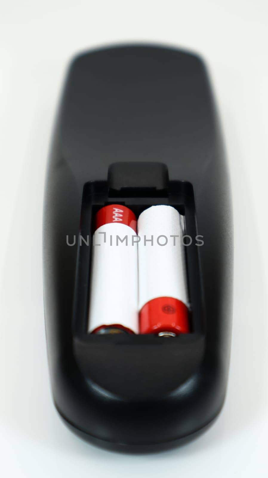 Black TV remote control with AAA alkaline batteries in red and white on a white background. Battery replacement, spare parts. Remote control battery compartment close-up. Vertical photography