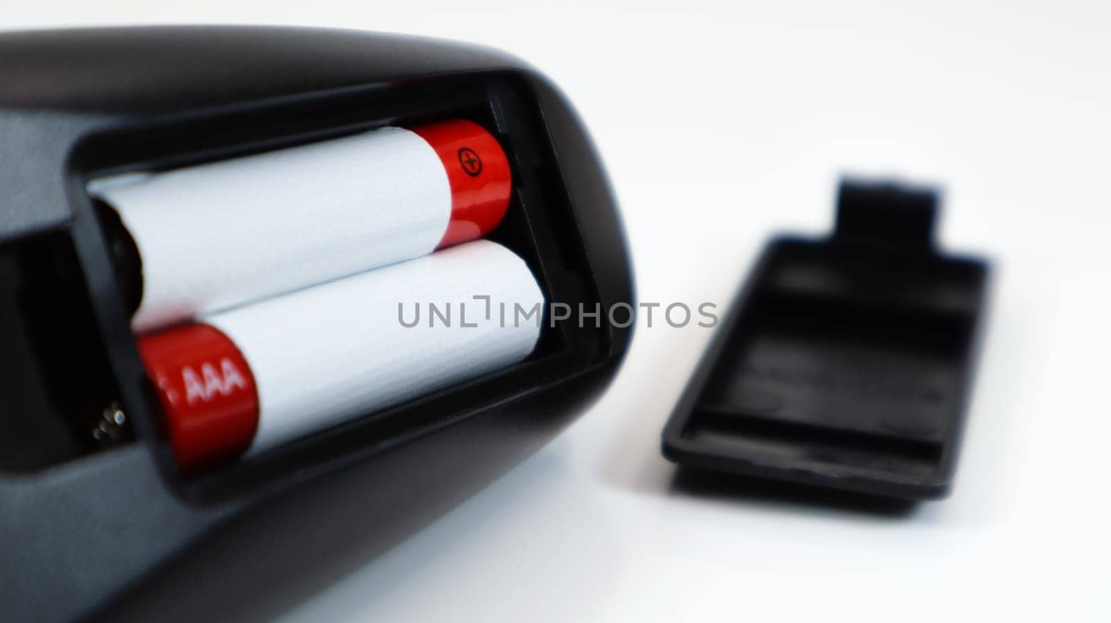 Black TV remote control with AAA alkaline batteries in red and white on a white background. Battery replacement, spare parts. Remote control battery compartment close-up