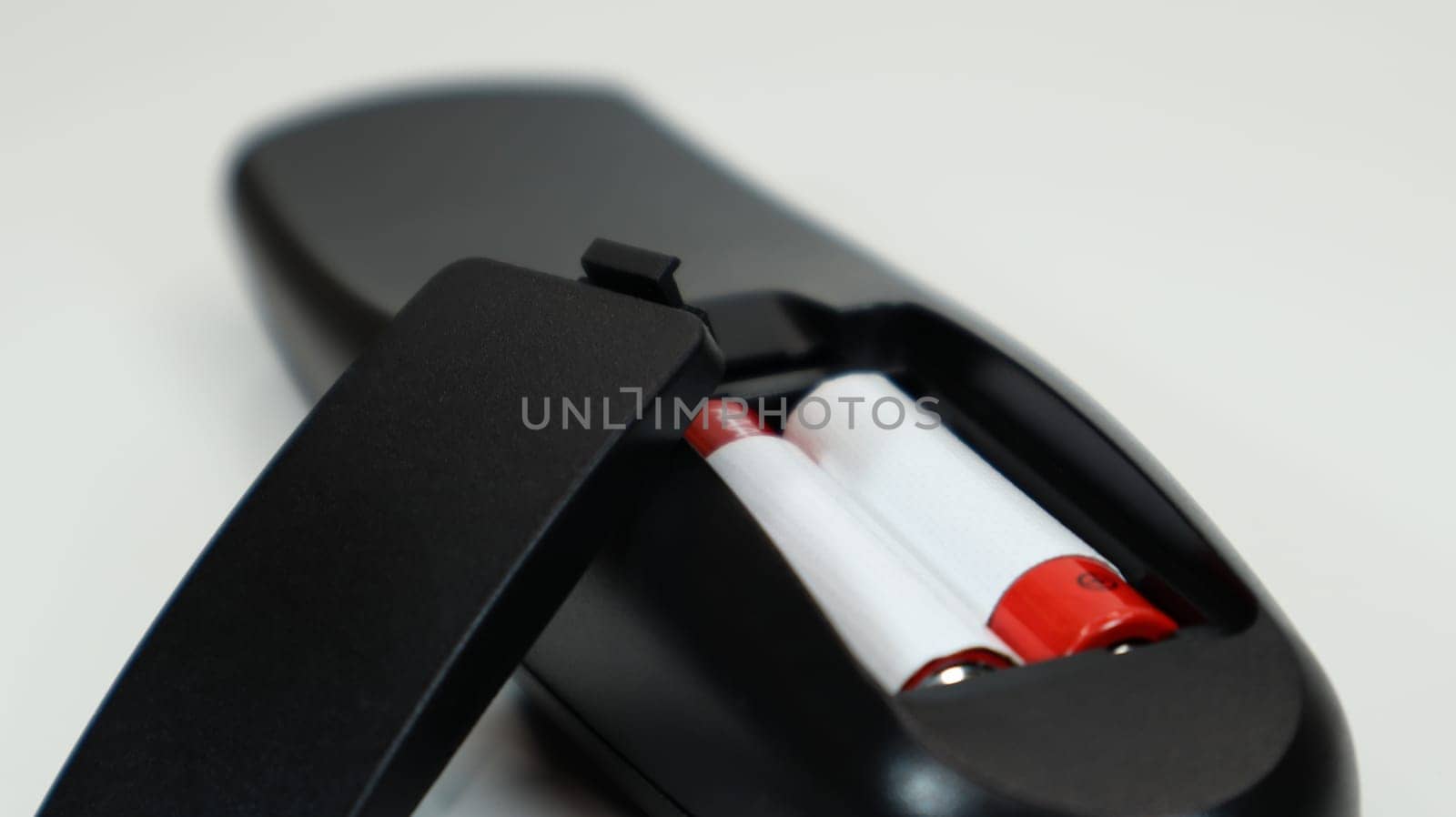 Black TV remote control with AAA alkaline batteries in red and white on a white background. Battery replacement, spare parts. Remote control battery compartment close-up