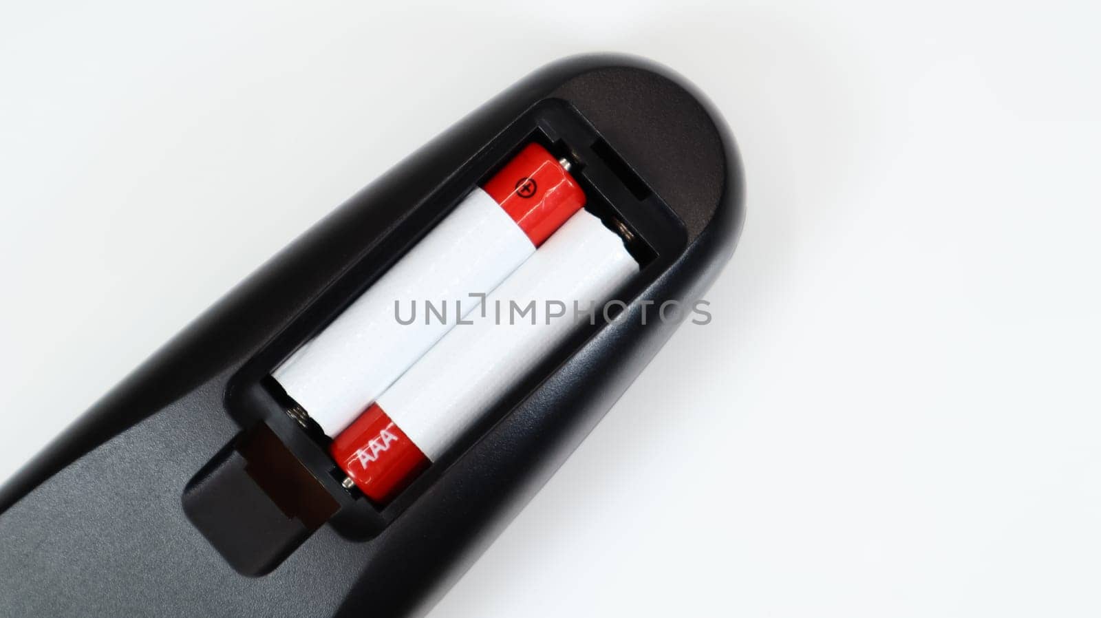 Black TV remote control with AAA alkaline batteries in red and white on a white background. Battery replacement, spare parts. Remote control battery compartment close-up. Copy space.