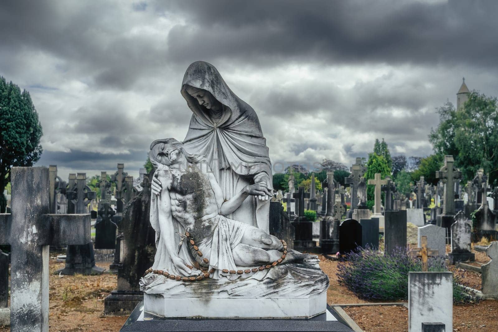 Dublin, Ireland, August 2019 Sculpture of Mary holding dying Jesus Christ, graves with Celtic crosses and sculptures in background, Glasnevin Cemetery
