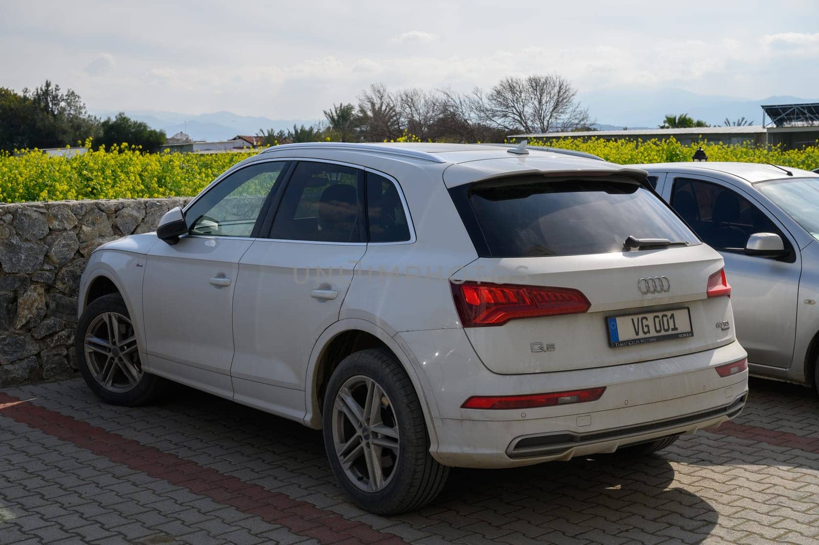 Gaziveren Cyprus -15.02.2025 A stylish white SUV rests near a stone wall, surrounded by vibrant greenery and a bright blue sky filled with clouds.