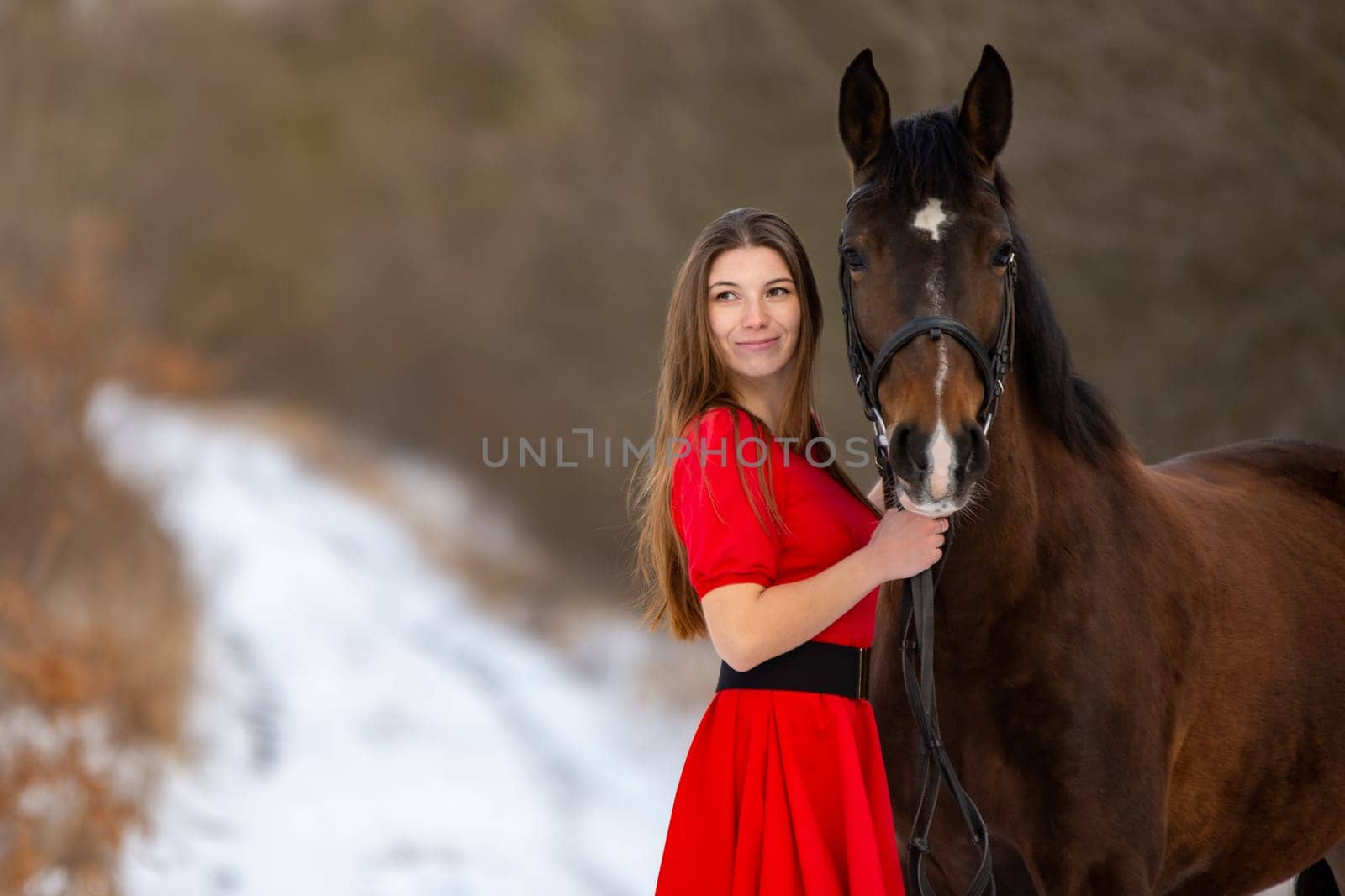 Portrait of a beautiful girl in a red dress standing with a horse on the background of a winter road