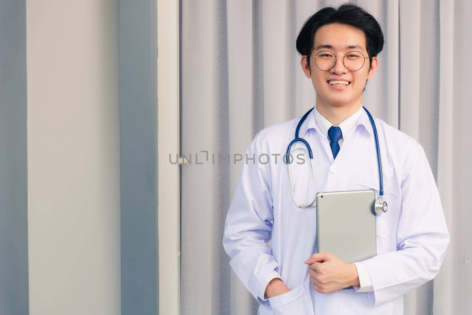 Portrait closeup of Happy Asian young doctor handsome man smiling in uniform and stethoscope neck strap holding smart digital tablet on hand and looking to the camera, healthcare medicine concept