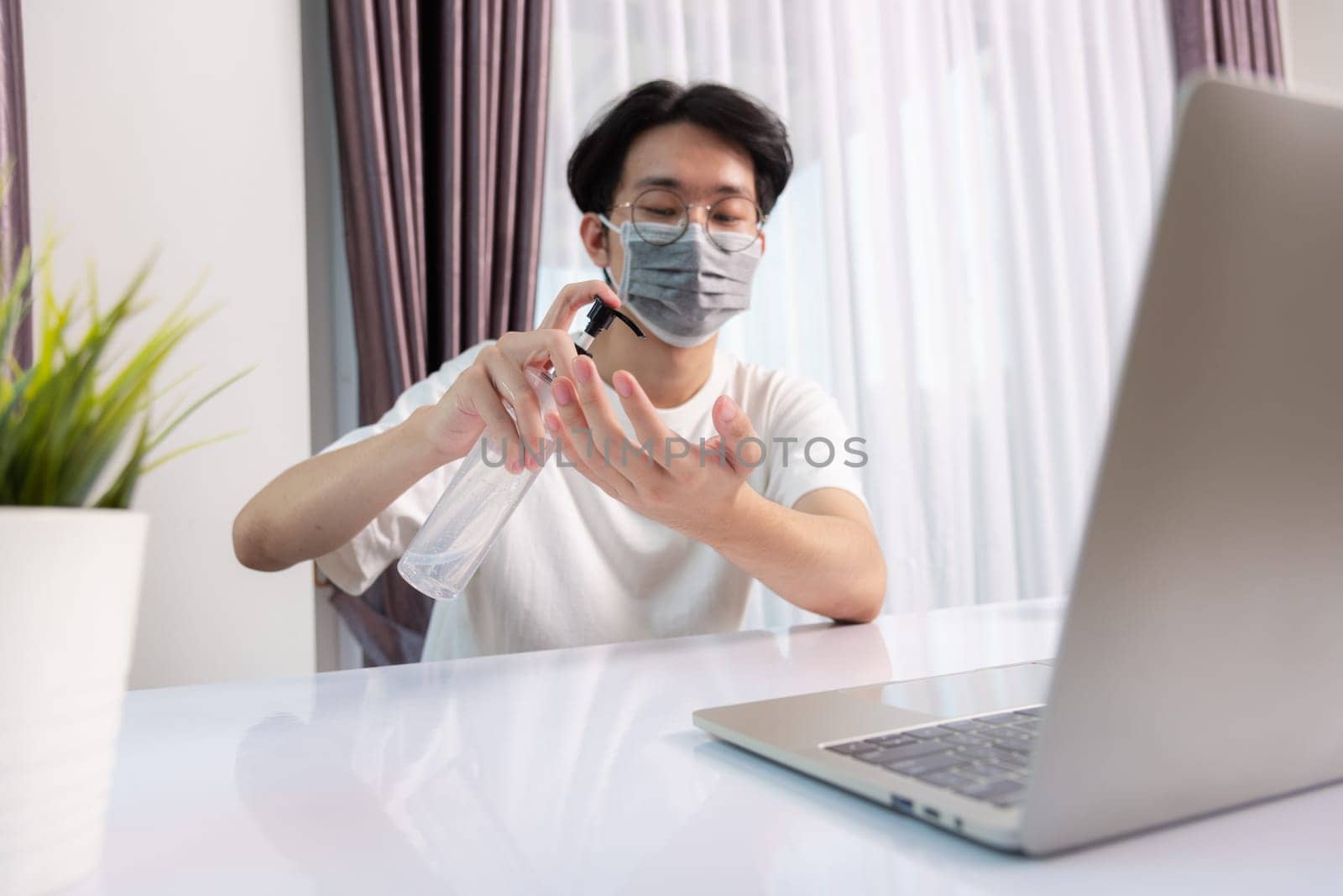 Asian business young handsome man glasses working from home office he quarantines disease coronavirus or COVID-19 wearing protective mask and cleaning hands with sanitizer gel on front laptop computer