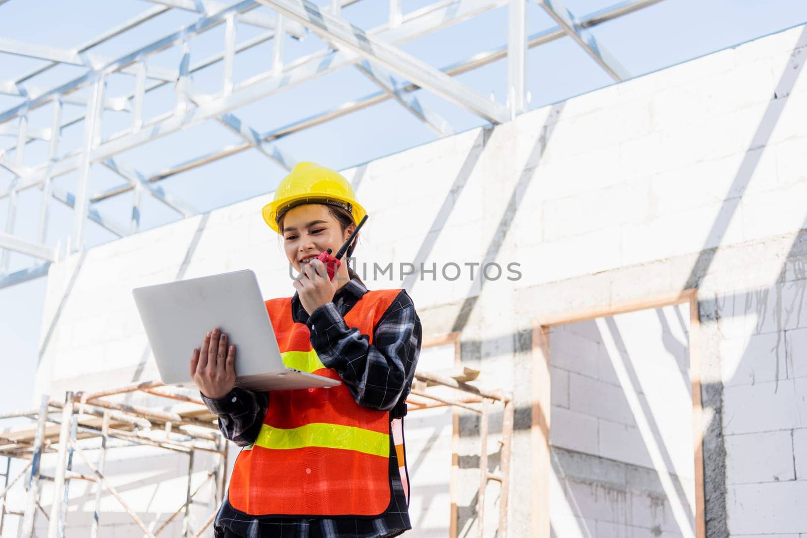 Asian engineer architect worker woman working at build construction site use laptop and talking with radio, engineering hold computer and radio discuss operate and control worker employee to building