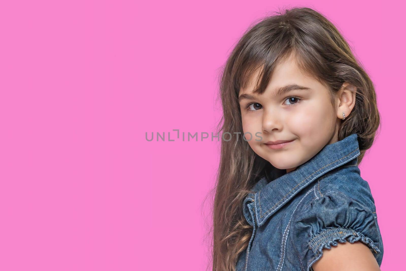 Smiling cute tanned long haired little girl wears  denim costume is posing on pink background.