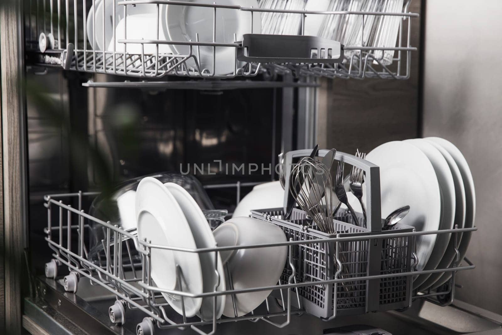 Front view of open automatic stainless steel built-in fully integrated top control dishwasher range machine with clean utensils, cutlery, glasses, dishes, plates inside in modern home kitchen