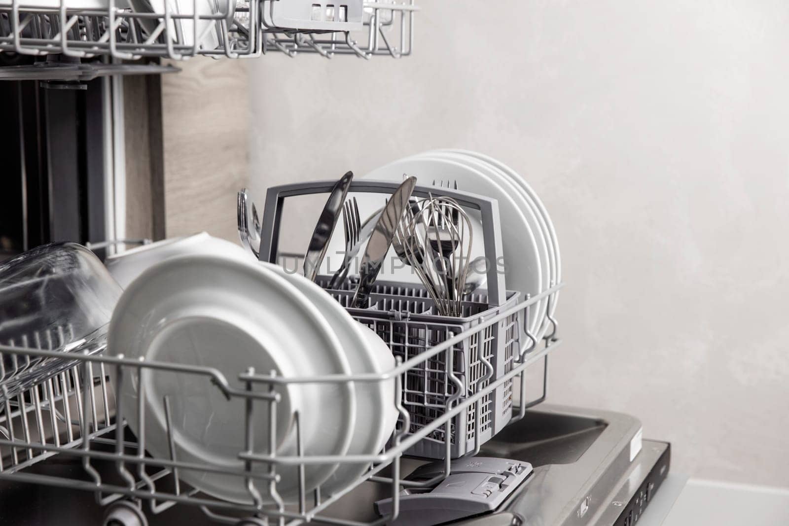 Front view of open automatic stainless steel built-in fully integrated top control dishwasher range machine with clean utensils, cutlery, glasses, dishes, plates inside in modern home kitchen