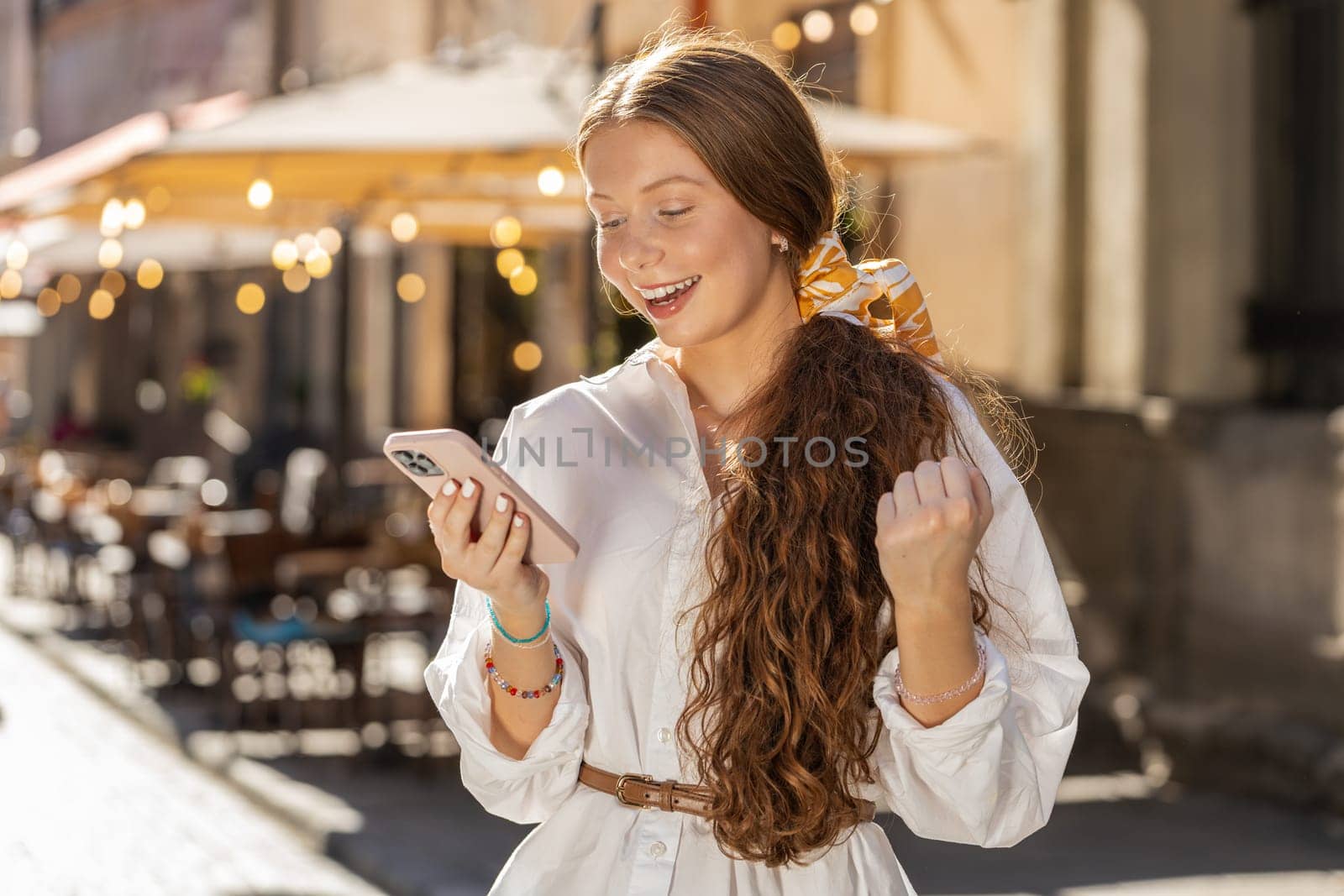 Teenager redhead girl use mobile smartphone celebrating win good message news, lottery jackpot victory, giveaway online outdoors. Happy young woman tourist walking in urban city street. Lifestyles