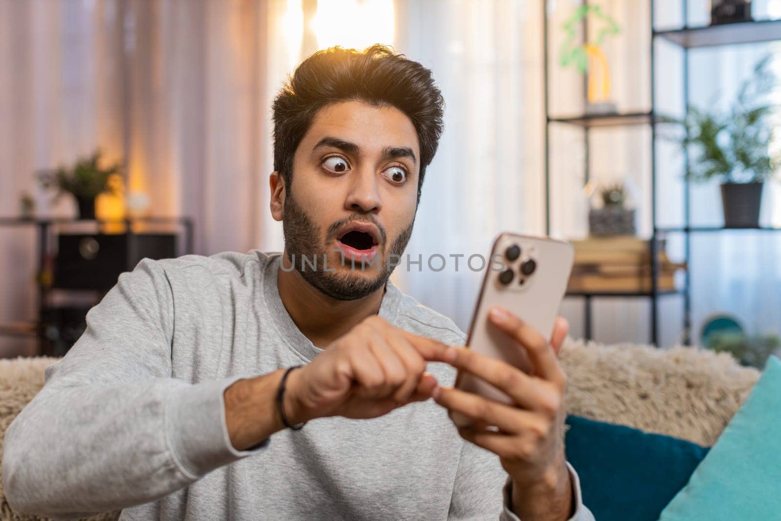 Happy young Indian man using smartphone scrolls through the news on smartphone reacting with surprise excitement wow. Hispanic male guy in casual clothes sitting on sofa couch in living room at home.