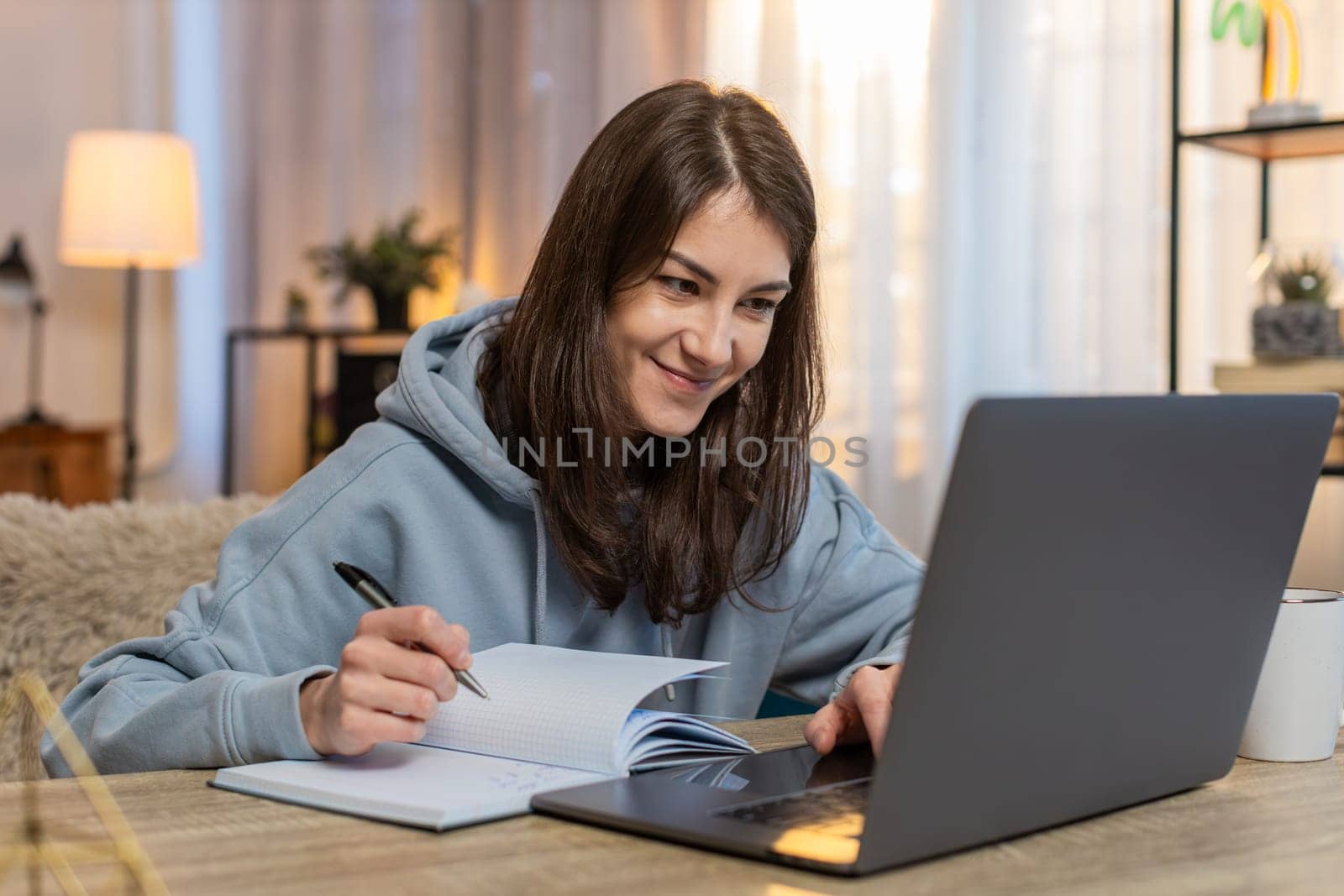 Young Caucasian woman watching online course on laptop sitting at table on sofa couch taking notes in notebook diary. Thoughtful female freelancer in casual clothes working from home in living room.