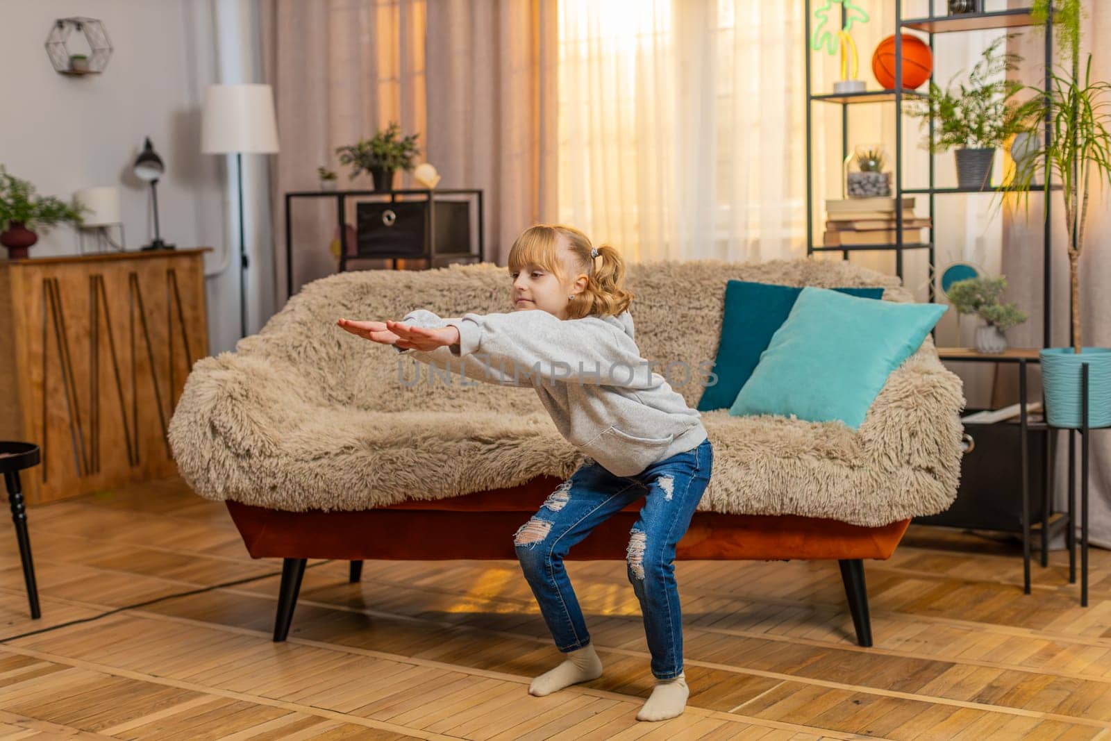 Young teenager girl stands at home performing simple squats and counting one, two, three with growing effort. Child kid sits on sofa finally collapses in laughter, feeling happy after a brief workout