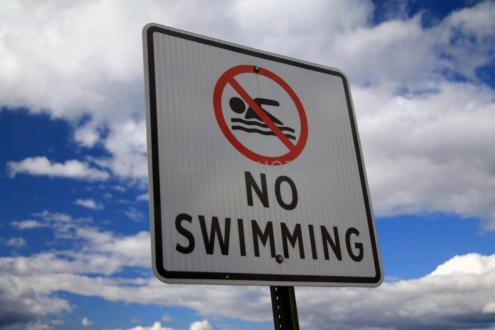 The No Swimming sign with the icon and a blue and white sky behind during a sunny day