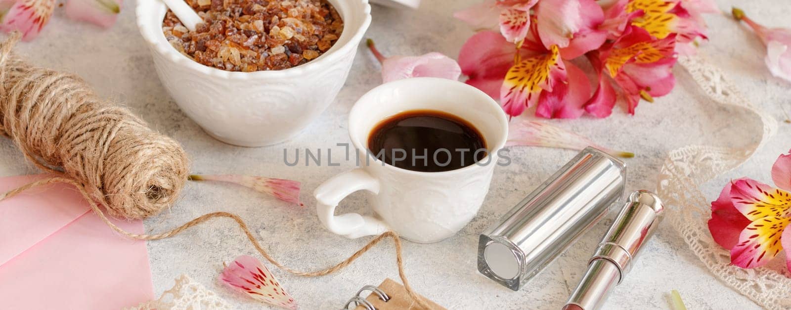 Coffee cup near pink flowers, lipstick and envelope on white table closeup. Romantic breakfast