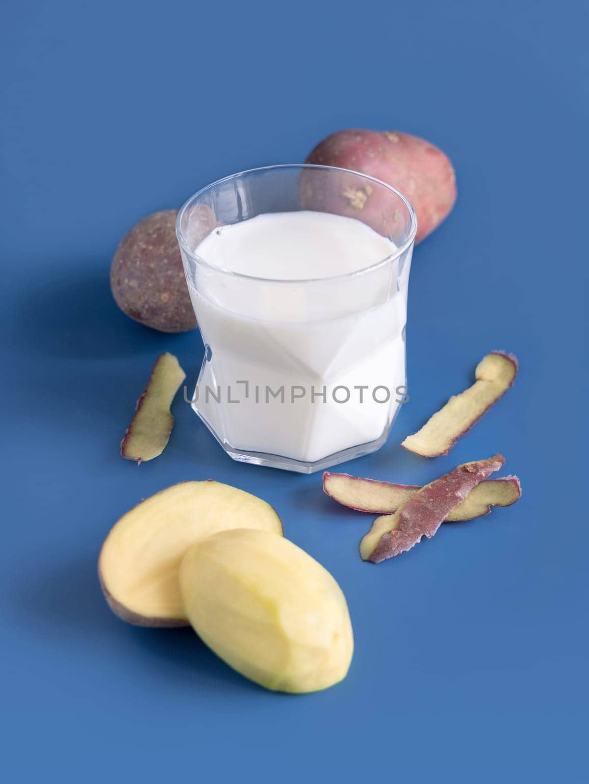 A transparent glass with vegetable potato milk on blue table near to potato tubers. Vegan plant based lactose free Milk close up. Healthy eating and Alternative food concept.