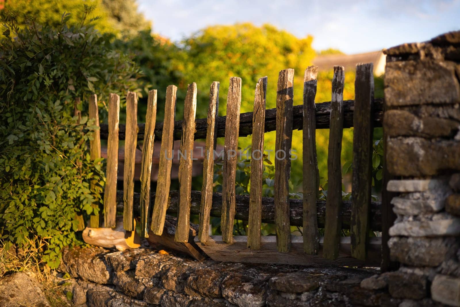 Old wooden picket fence built into a wall from stones. Sunset over an aged timber barrier protecting privacy of home on a village. Hardwood brown material.