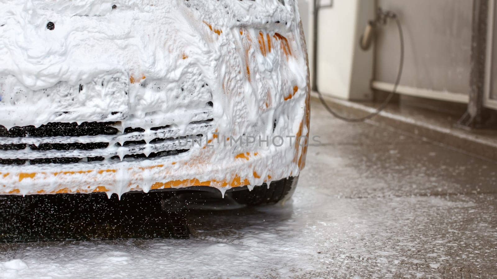 Front bumper of yellow car being washed in carwash, completely covered in white shampoo and foam, some dropping to the ground.
