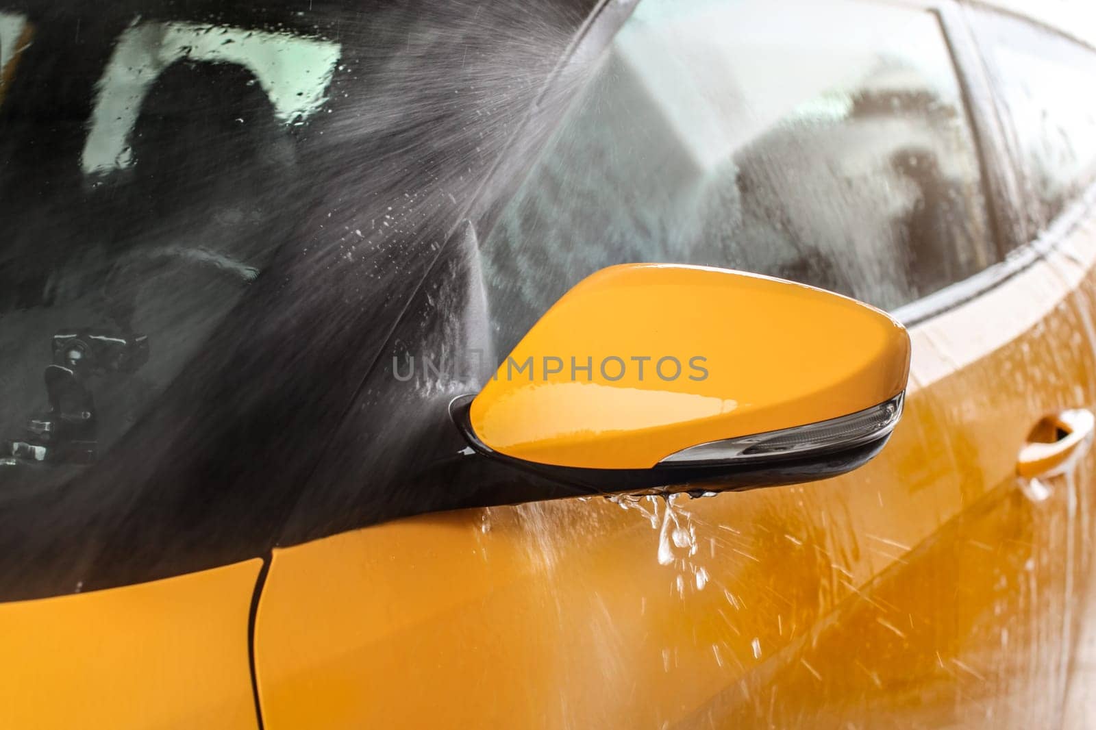 Side mirror of yellow car being washed in self service carwash, water spraying with high pressure, drops flying around.