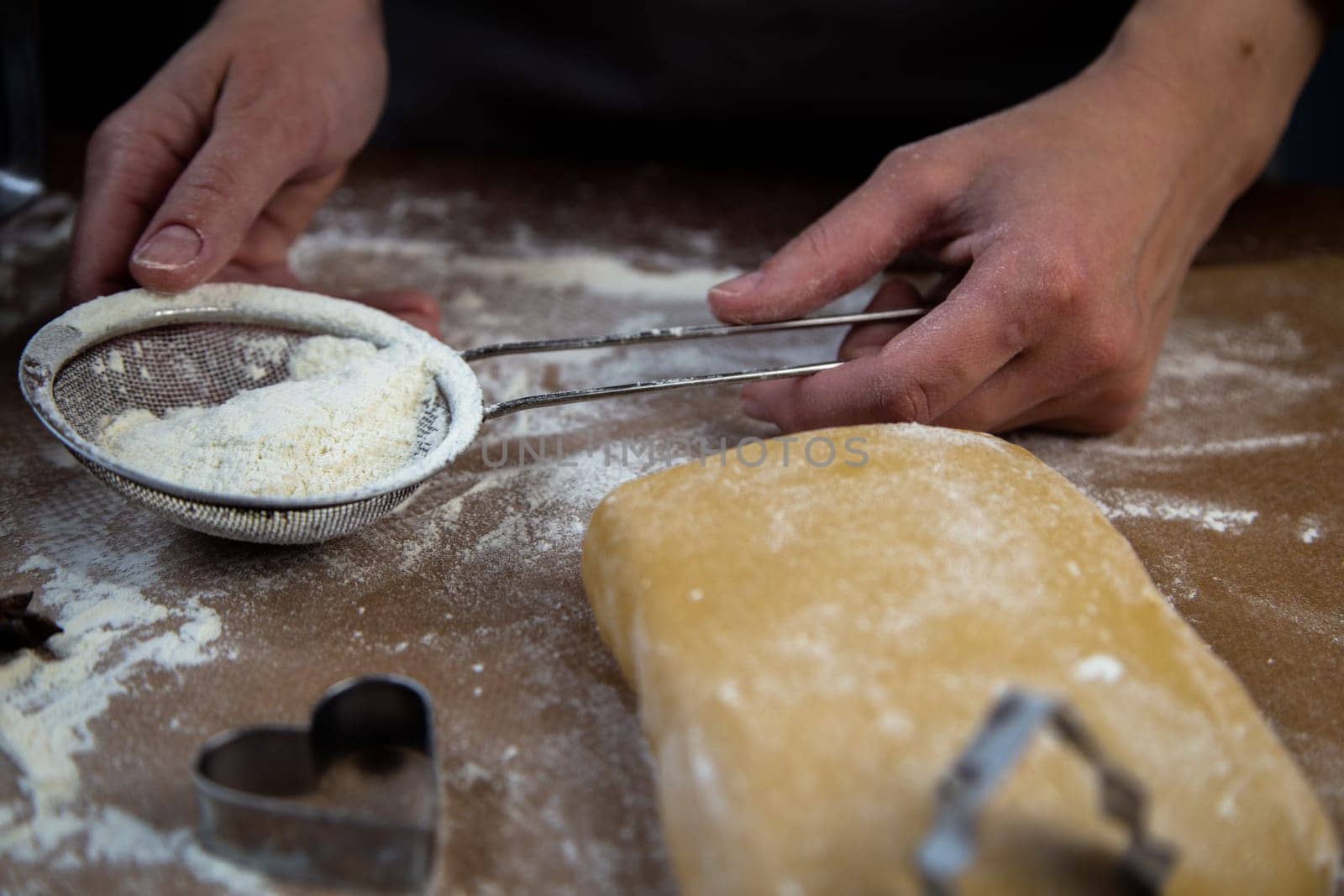 The chef's hands hold a sieve with flour on the background of parchment, flour, dough and cookie molds. High quality photo