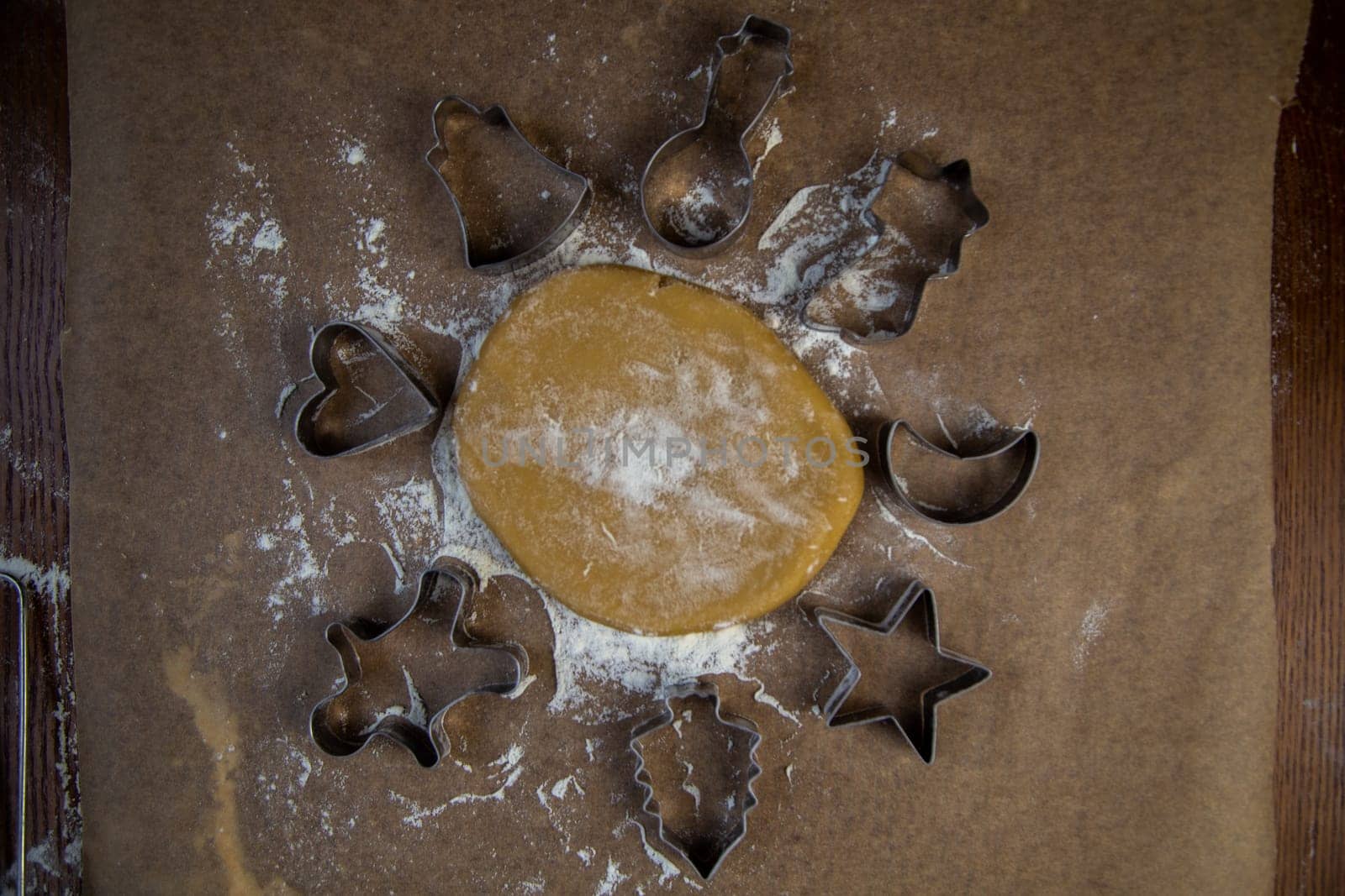 There is a dough on the parchment, around which there are cookie cutters of different shapes, all sprinkled with flour. High quality photo