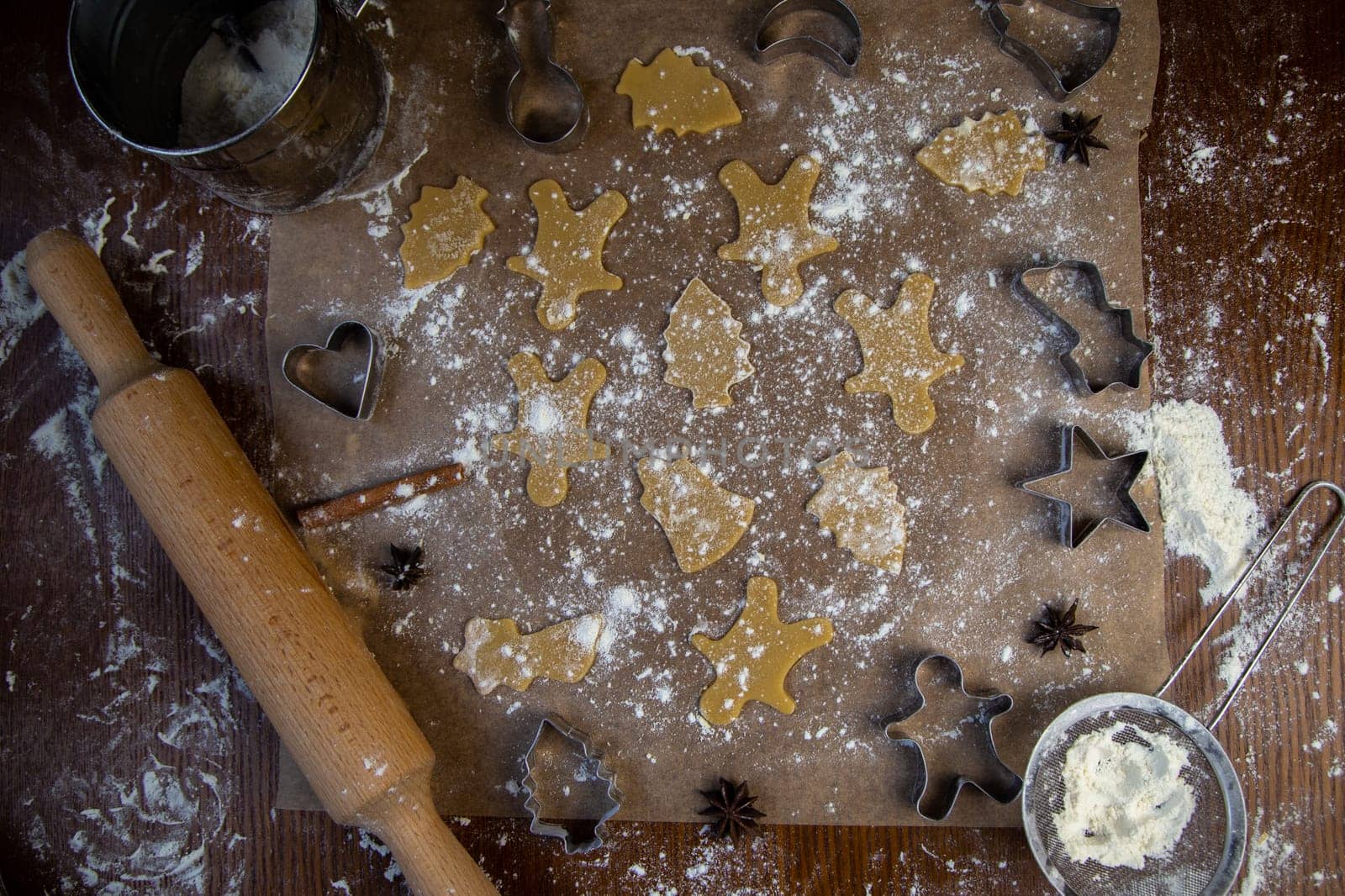 There are ready-made cookies of different shapes on the parchment, a rolling pin, cookie cutters and a sieve with flour are lying nearby, flour is scattered everywhere. High quality photo