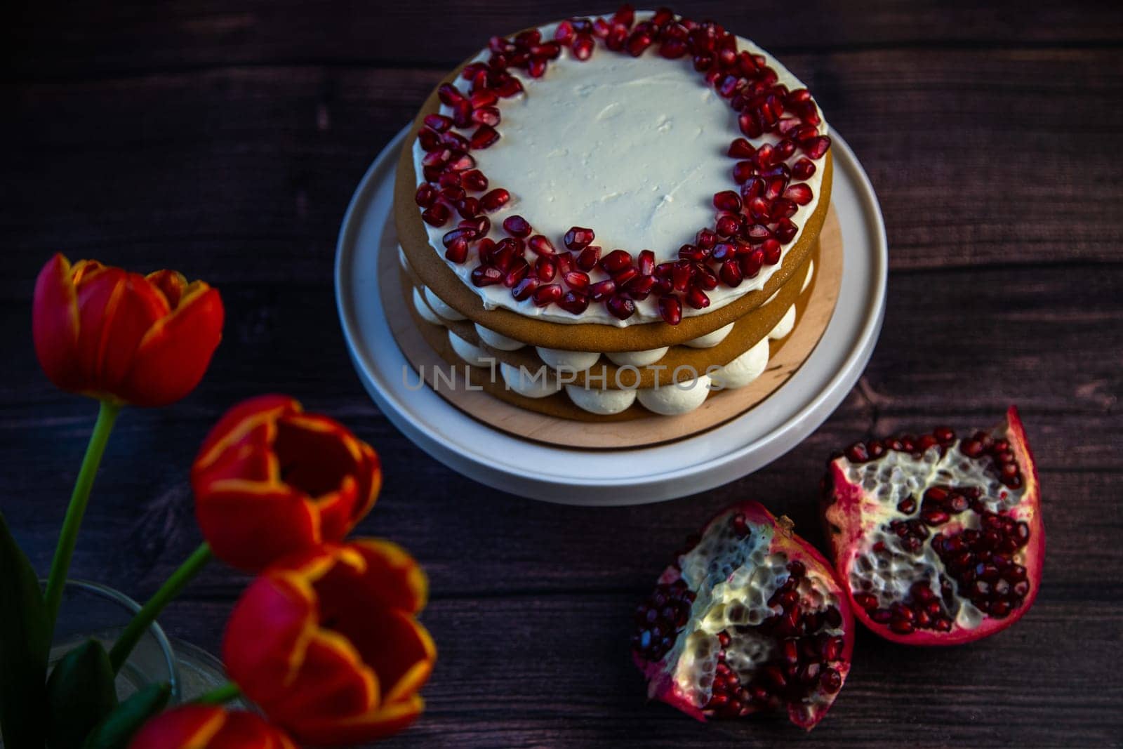 A cake of three layers consisting of cakes and white cream, decorated with pomegranate on top in a circle, stands on a dark background, next to tulips and pomegranates. High quality photo