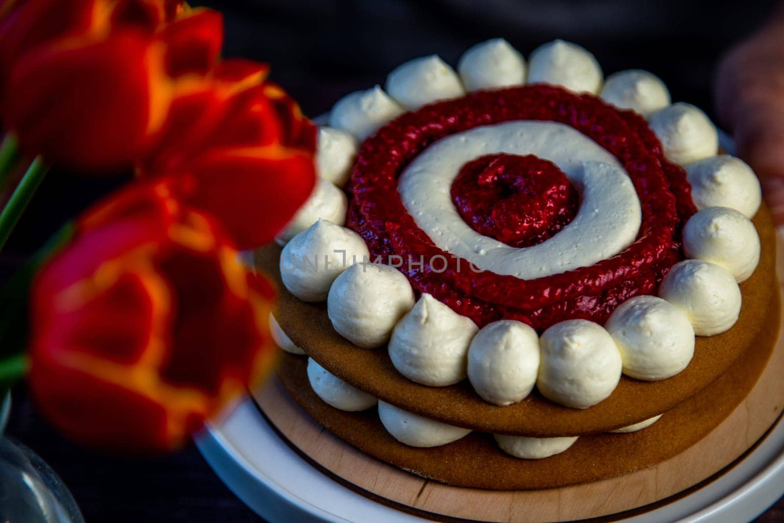 The baked cake cake lies on a wooden substrate and on a white stand, the cake is decorated with white cream and raspberry jam, in a circle, circles alternate, a bouquet of red tulips on a dark background is visible nearby. High quality photo
