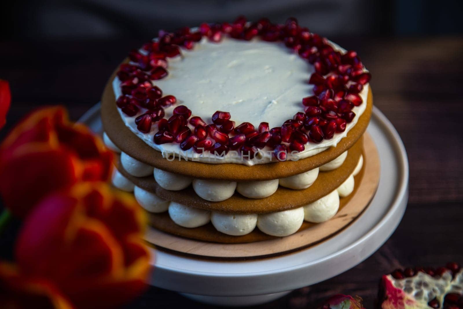 A cake of three layers consisting of cakes and white cream, decorated with pomegranate on top in a circle, stands on a dark background, next to tulips and pomegranates. High quality photo