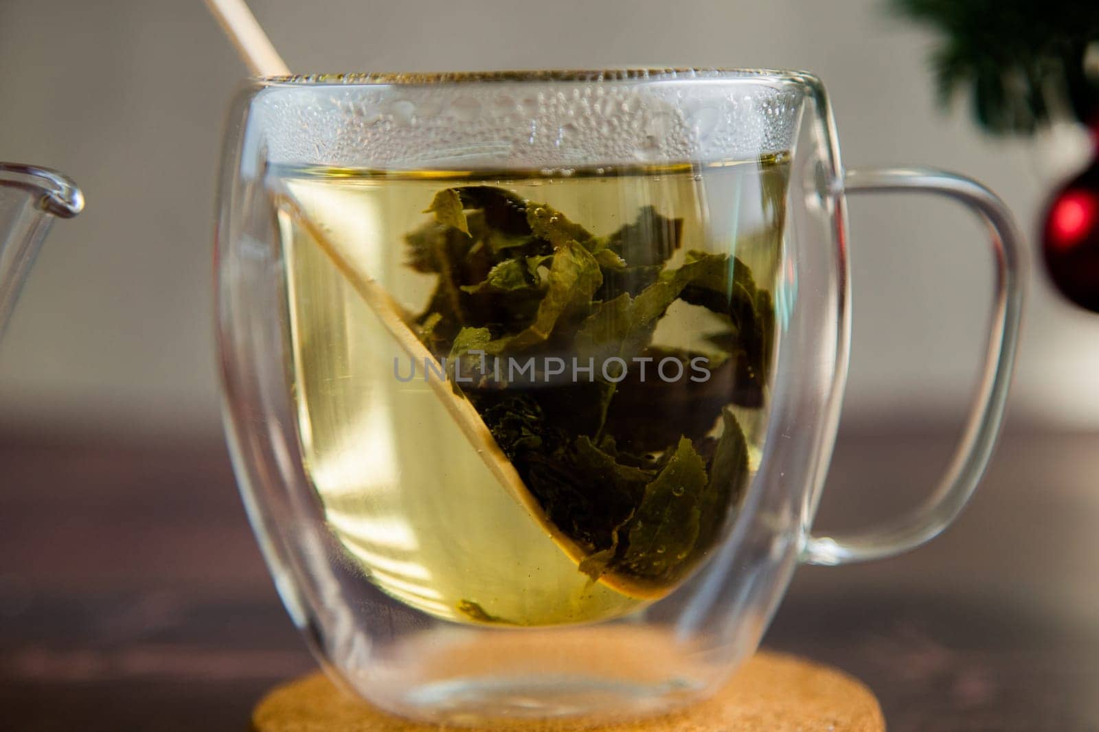 A transparent cup of tea stands on a wooden stand, green tea leaves and a wooden spoon float in the mug. High quality photo